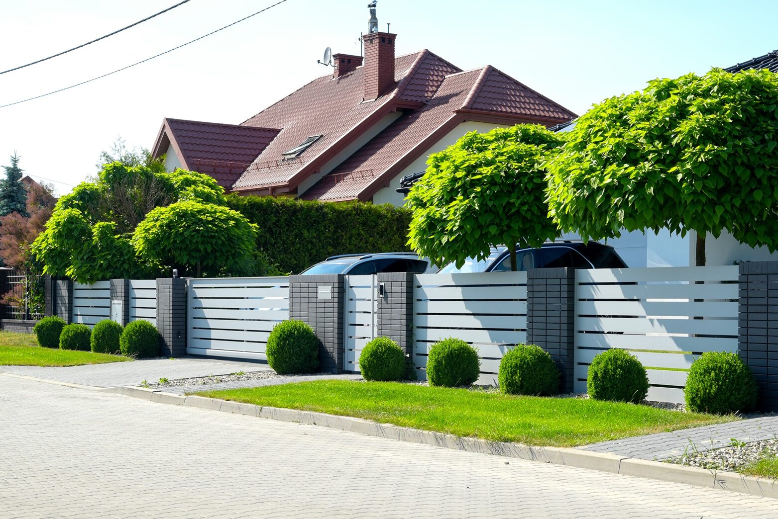 Na obrazku jest przykład pięknego, biało - antracytowego ogrodzenia aluminiowego, idealnie zharmonizowanego z bryłą budynku i roślinnością.