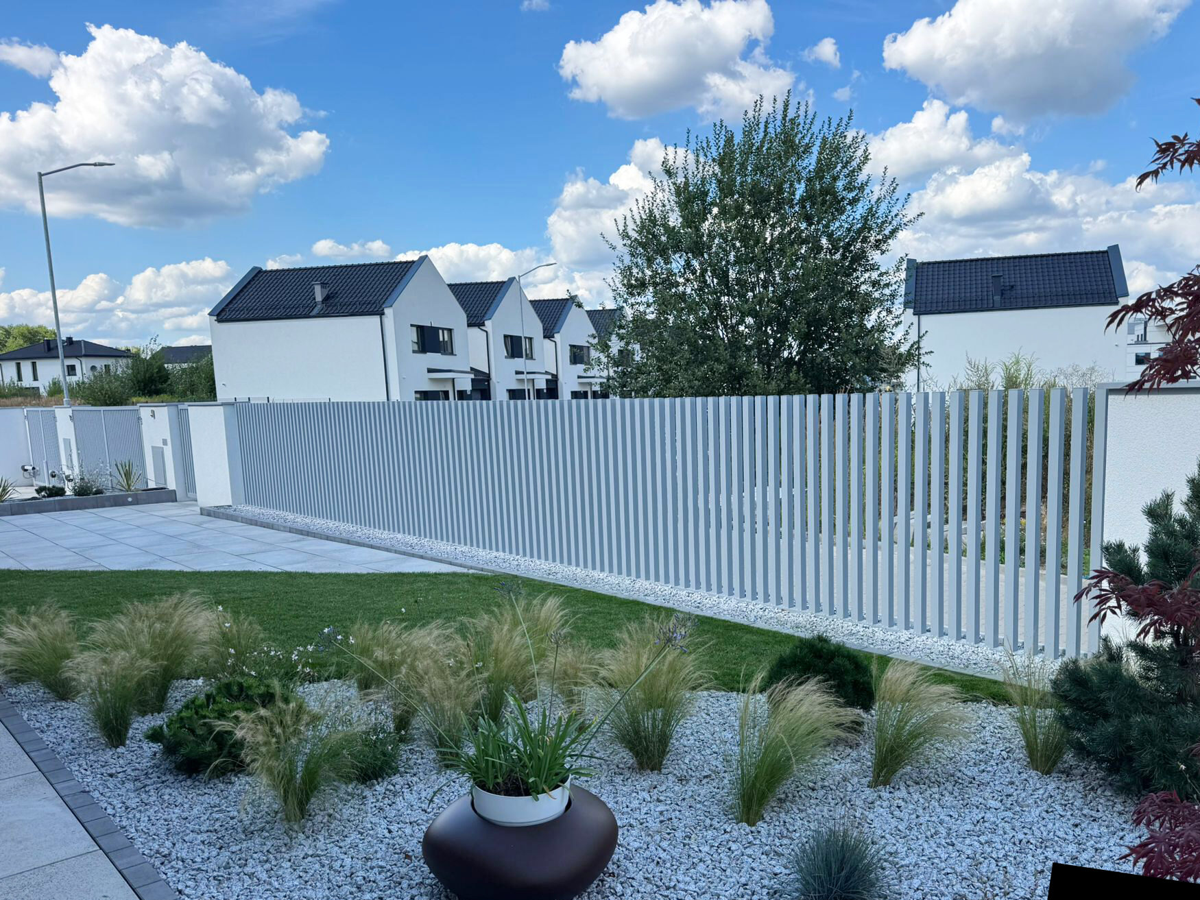 Przykład minimalistycznego, białego ogrodzenia aluminiowego palisadowego pionowego, zaskakującego estetyką. Pięknie komponuje się z białym żwirem i trawnikiem w ogrodzie.