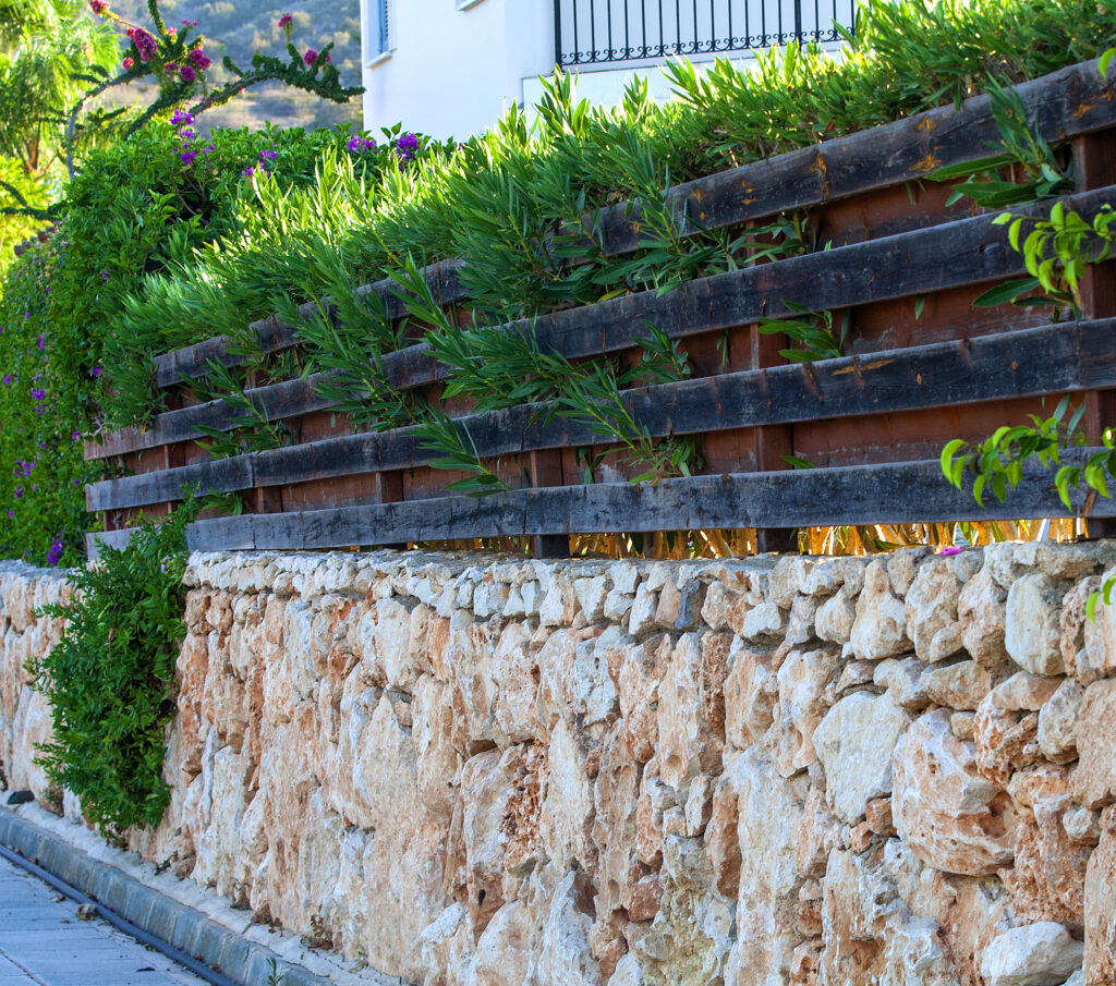 Przykład połączenia 3 różnych ogrodzeń w jedno - na dole kamienne, wyżej drewniane, wzmocnione aluminiowymi palisadami, i z żywopłotem.